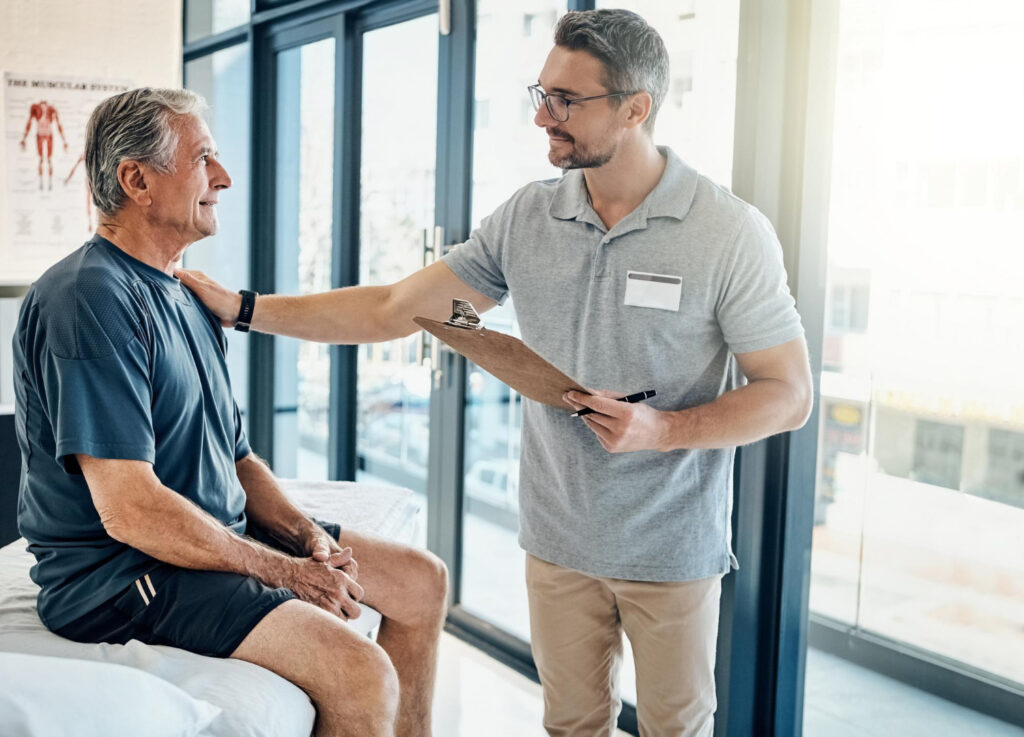 Physiotherapist with a clipboard reassuring an older male patient during a consultation